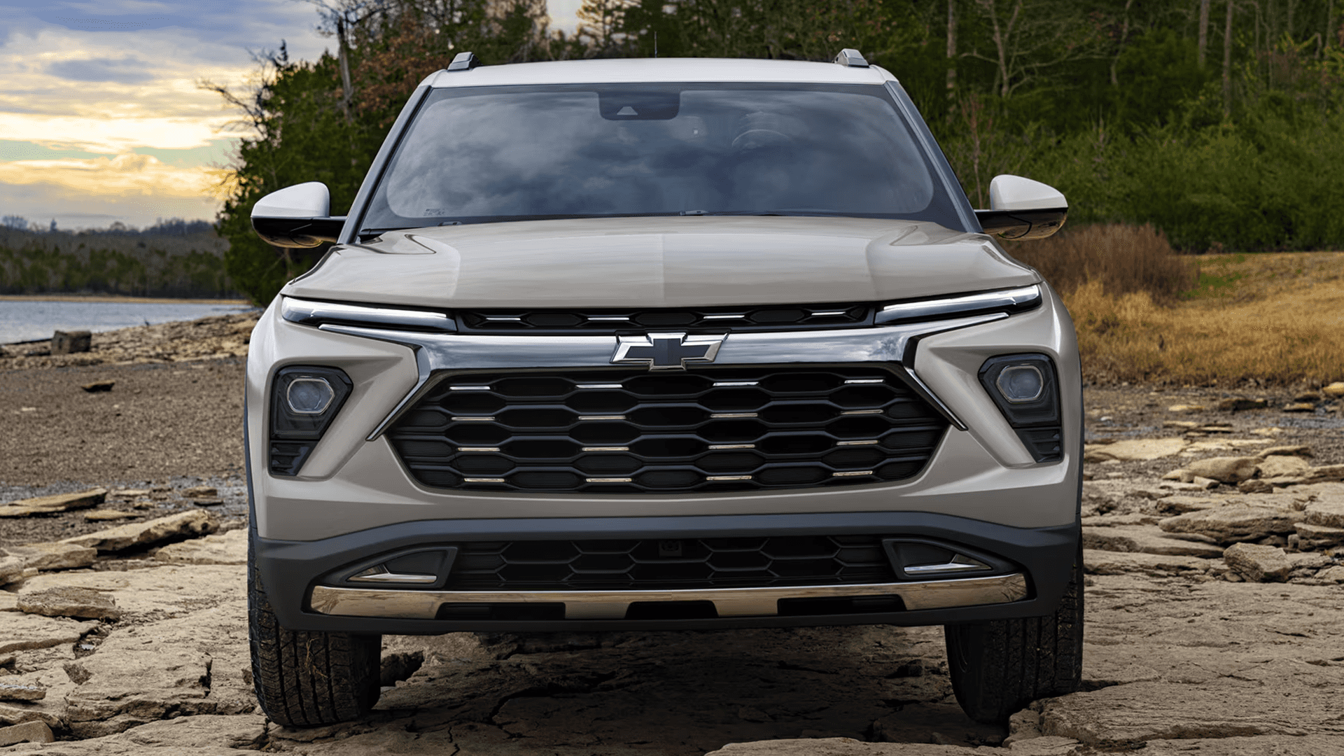 Front view of a tan 2026 Chevrolet Trailblazer showing the wide grille with Chevrolet bowtie badge, slim upper lighting, and lower headlamp housings, photographed on a rocky shoreline with trees and a cloudy sunset sky behind it.