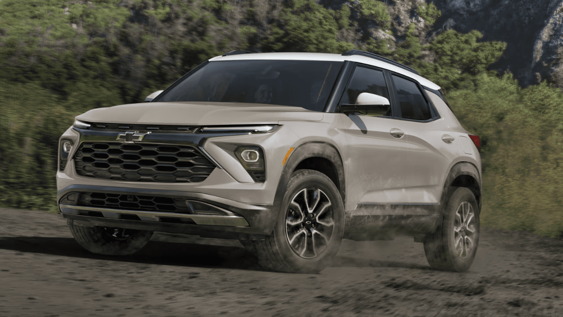 A tan 2026 Chevrolet Trailblazer drives on a dusty dirt road, kicking up dust around the wheels, shown in a three-quarter front angle with the Chevrolet bowtie on the grille and a wooded hillside in the background.