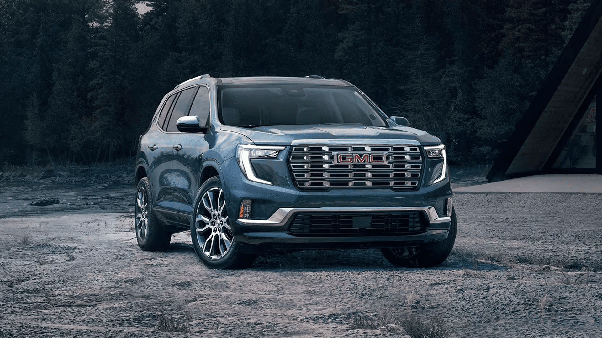 Blue 2026 GMC Acadia parked on a gravel surface in front three quarter view, highlighting the wide grille, C shaped LED running lights, sculpted hood, and large polished wheels near a wooded backdrop.