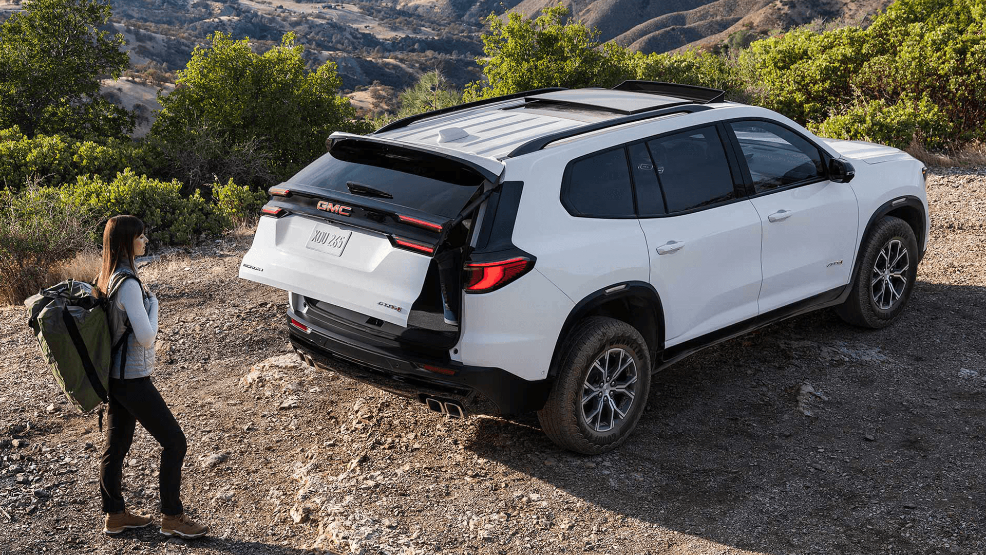 Rear three quarter view of a white 2026 GMC Acadia with the liftgate open on a hillside trail, showing cargo access, roof rails, rear LED lighting, dual exhaust outlets, and a hiker standing nearby.