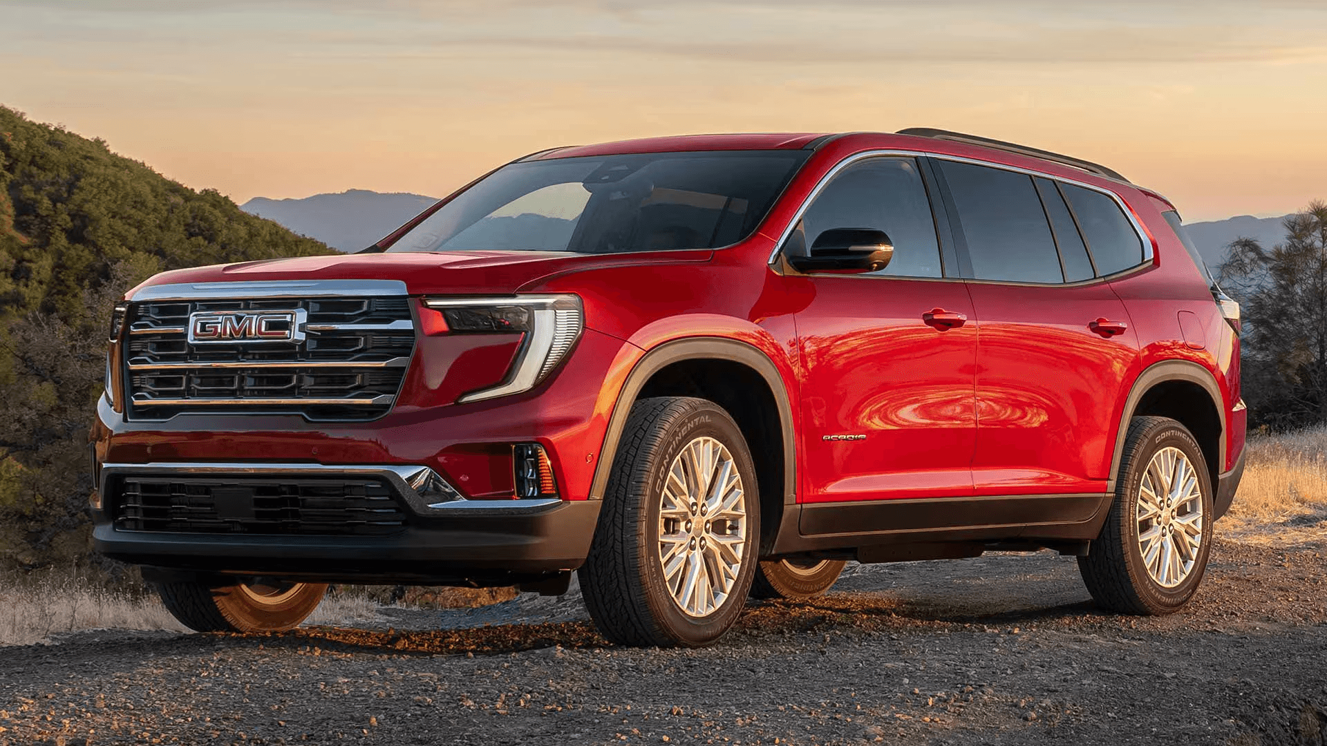 Red 2026 GMC Acadia shown in a front three quarter view at sunset, with a bold GMC grille, LED signature lighting, and bright multi spoke alloy wheels on a gravel overlook.