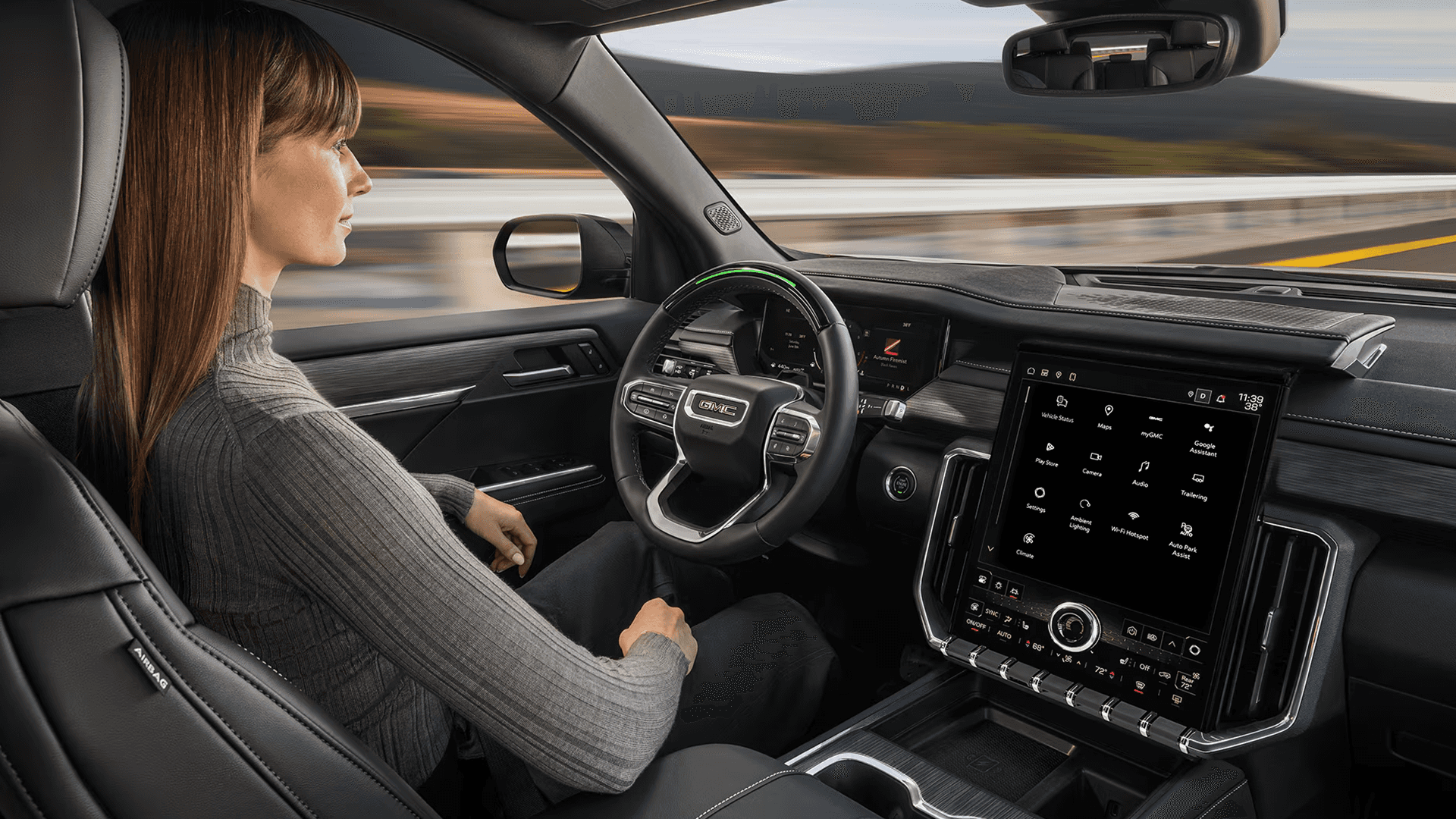 Front seat cabin view of a driver in a 2026 GMC Acadia with hands off the wheel, highlighting a green light bar on the steering wheel and the large portrait touchscreen, suggesting hands free driver assistance use.