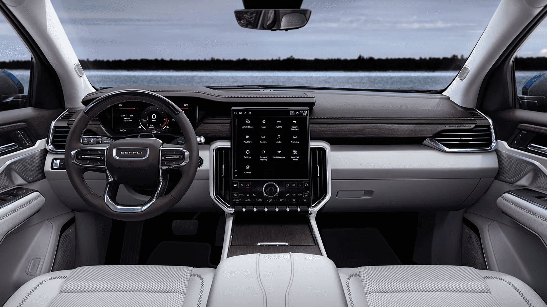 Full width view of the 2026 GMC Acadia Denali interior featuring light seats, wood tone dash trim, a digital gauge cluster, and a large portrait center touchscreen, with a lake visible through the windshield.