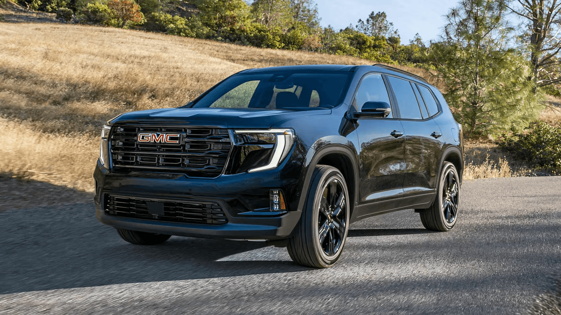 Dark blue GMC SUV driving on a paved road with grassy hills and trees in the background, captured from a front three quarter angle to highlight the grille, LED lighting, and black wheels.