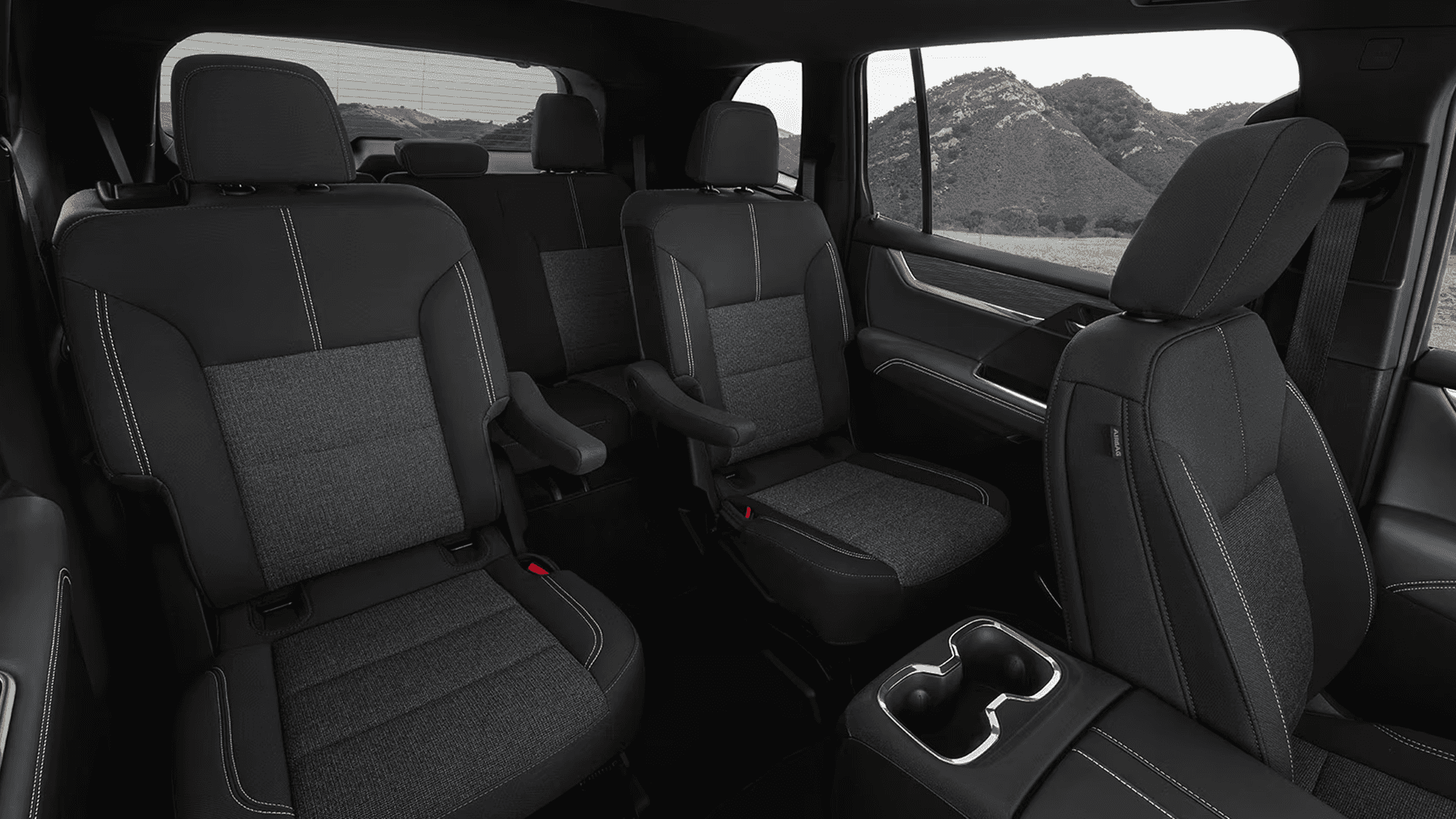 Interior view of a GMC SUV showing second row captain’s chairs and third row seating in black upholstery with contrast stitching, with a center armrest and cupholders visible and mountains outside the side window.