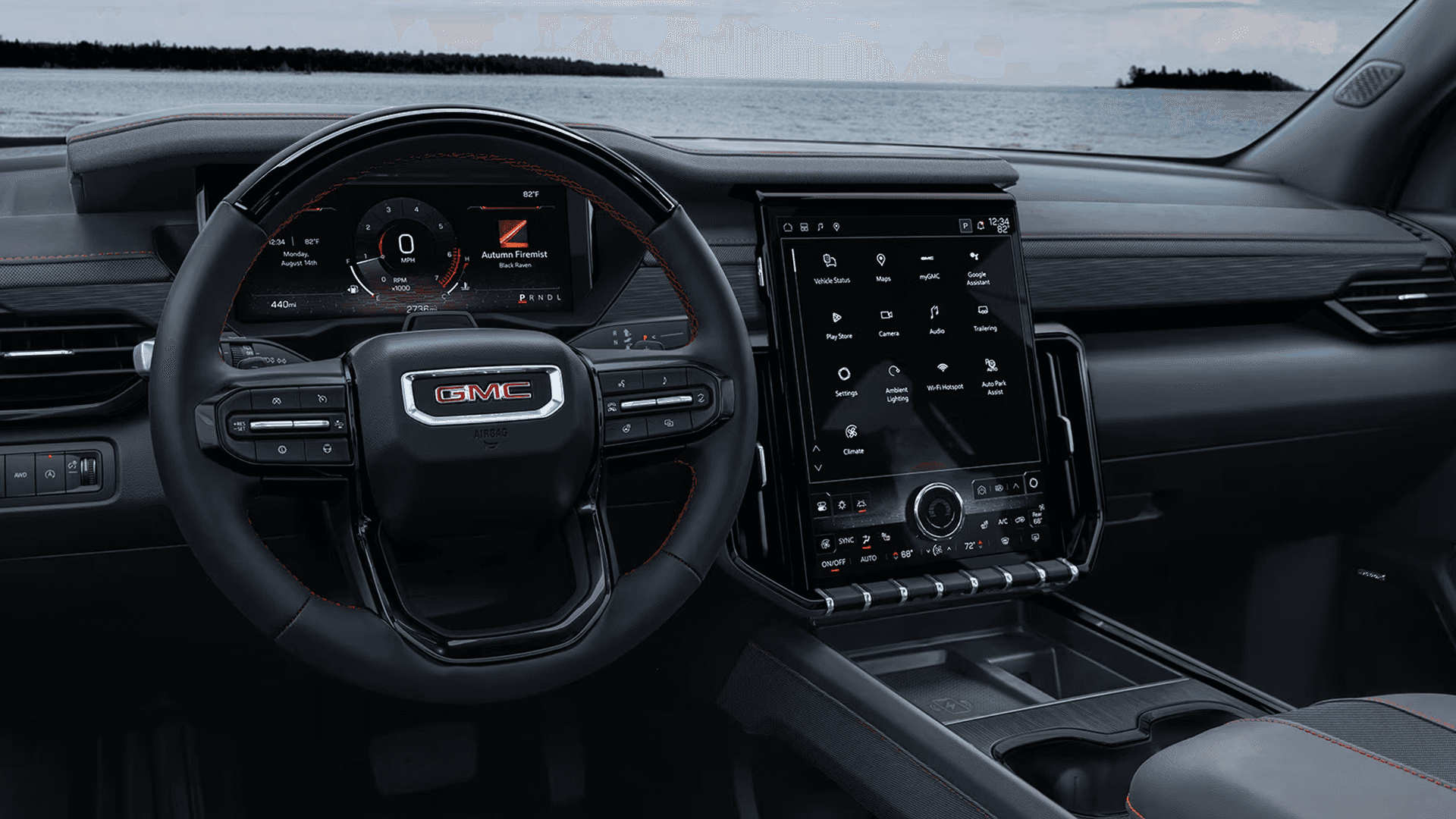 Driver cockpit view showing the GMC steering wheel, digital instrument cluster, and a large vertical touchscreen displaying app icons, with the center console and cupholders in the foreground and a shoreline visible through the windshield.