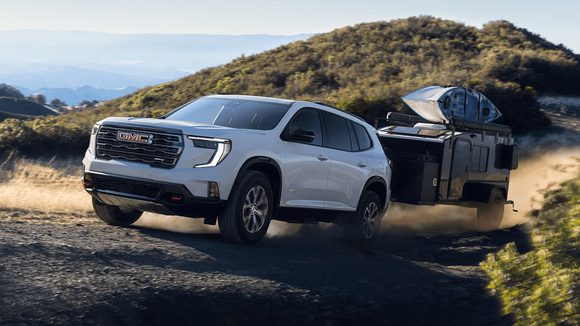 White GMC SUV towing an off road camper trailer on a dirt road, kicking up dust, with a kayak mounted on the trailer rack and hills and mountains in the background.