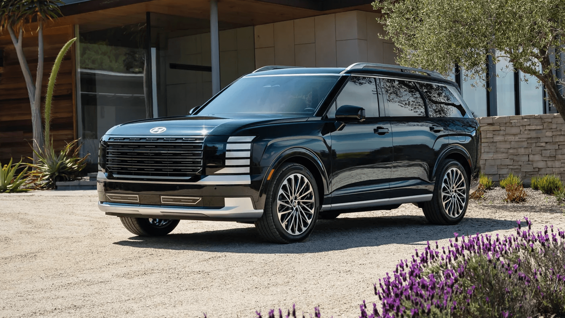 Front three-quarter view of a dark green 2026 Hyundai Palisade parked outside a modern home, showing the bold grille, stacked LED lighting signature, sculpted body lines, silver lower accents, tinted windows, and multi-spoke wheels beside purple flowers.