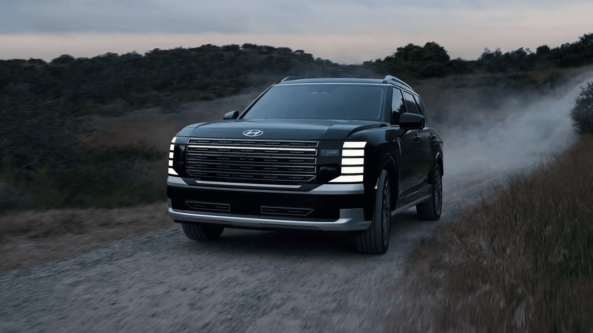 Front three-quarter view of a dark green 2026 Hyundai Palisade driving on a gravel road at dusk, with the stacked LED running lights illuminated and dust trailing behind, showing the wide grille, roof rails, and silver lower bumper trim.