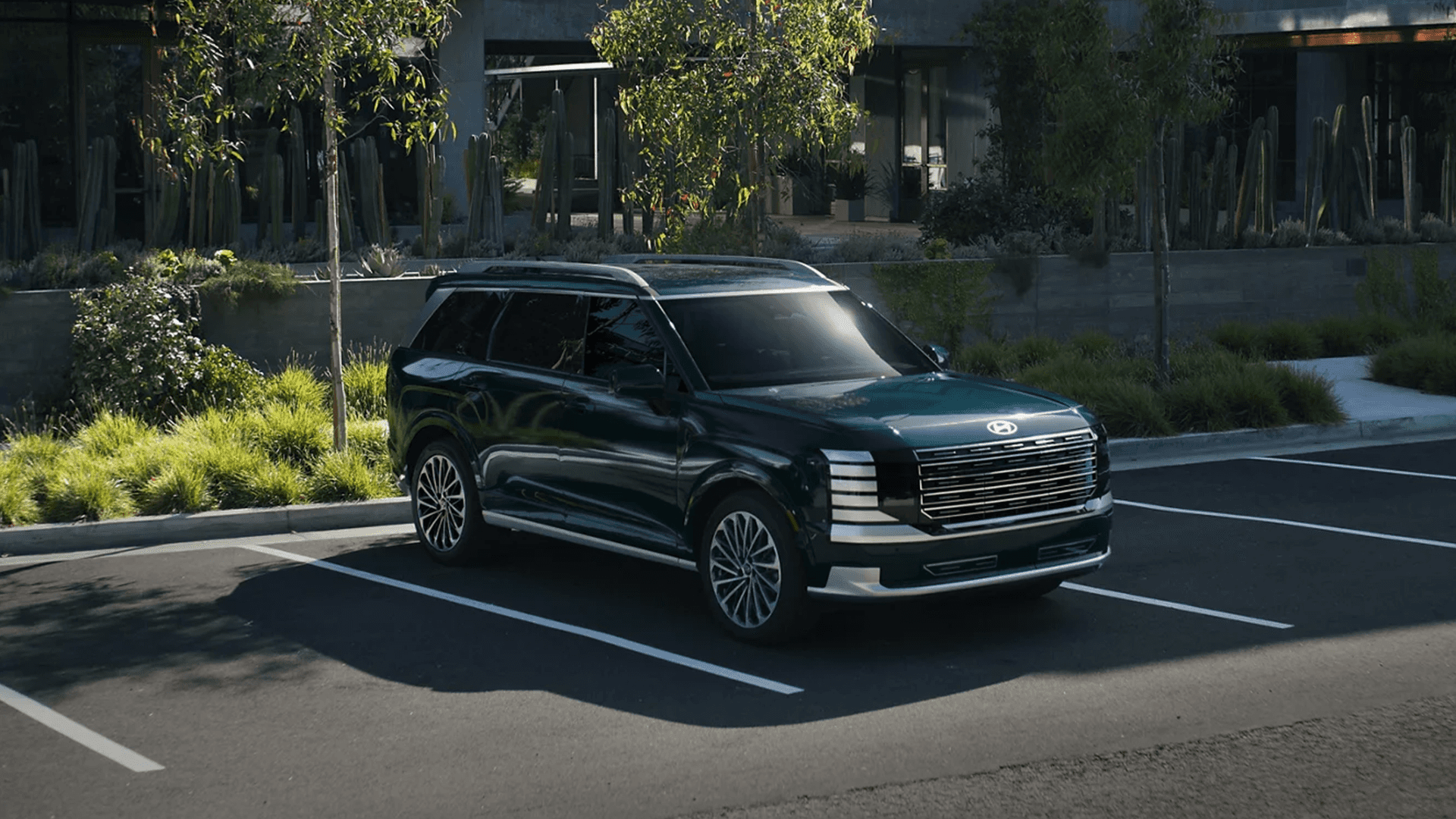 Dark green 2026 Hyundai Palisade parked diagonally in an empty parking space near a modern building and landscaping, highlighting the roof rails, long SUV profile, large wheels, and upright front design.