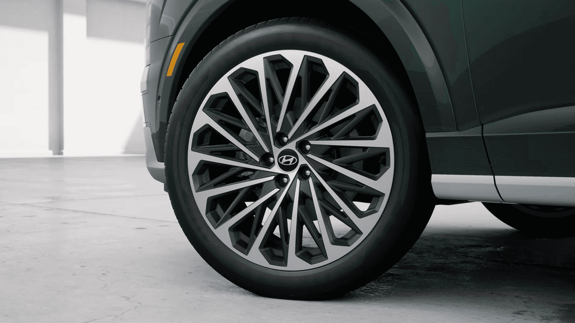 Close-up of a front wheel on a dark green 2026 Hyundai Palisade, showing a large two-tone multi-spoke alloy wheel with the Hyundai center cap, black tire sidewall, and lower body cladding near the rocker panel.