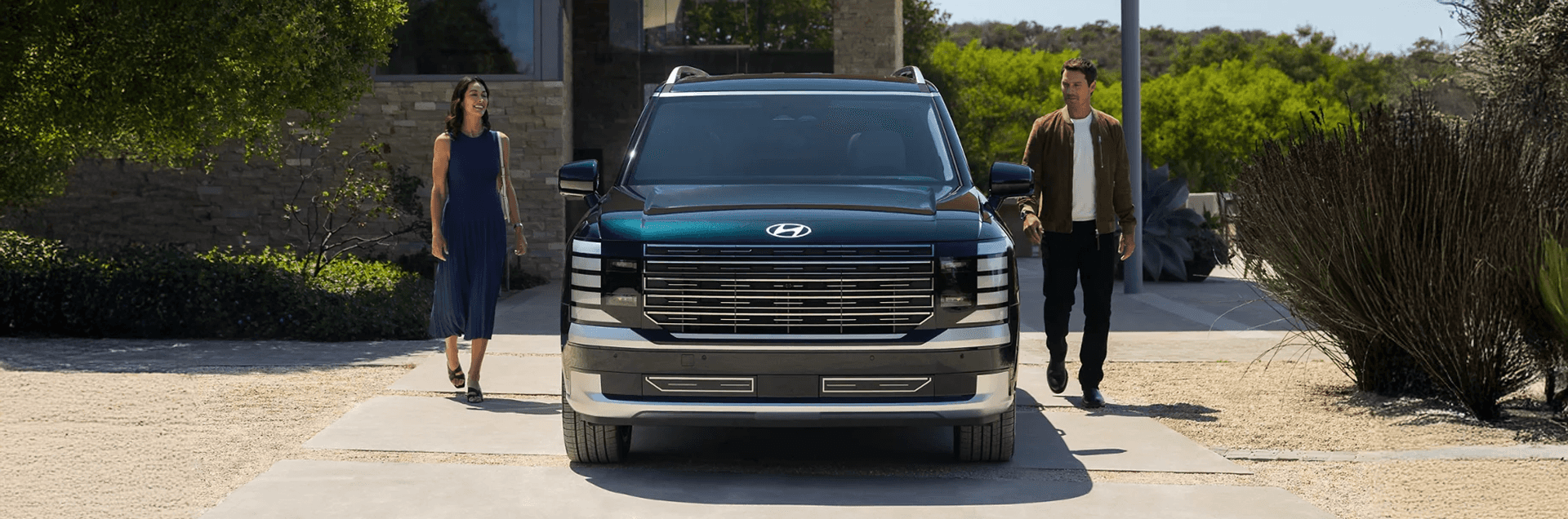 Front view of a dark green 2026 Hyundai Palisade parked on a driveway, showing the wide grille, stacked horizontal LED running lights, silver lower trim, and roof rails, with two people walking on either side.