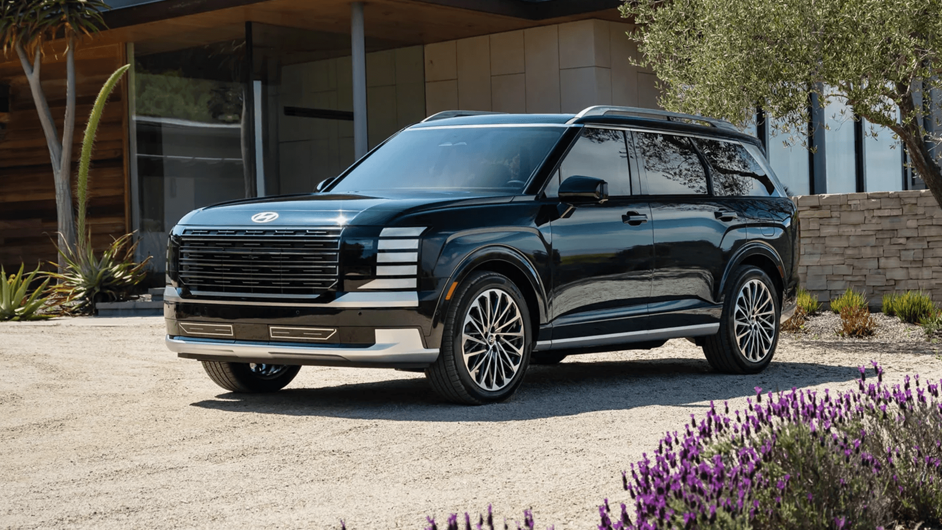 Side front three quarter view of a dark green 2026 Hyundai Palisade in a modern courtyard, highlighting the squared body lines, black window trim, roof rails, large grille, vertical LED lighting elements, and silver lower cladding.
