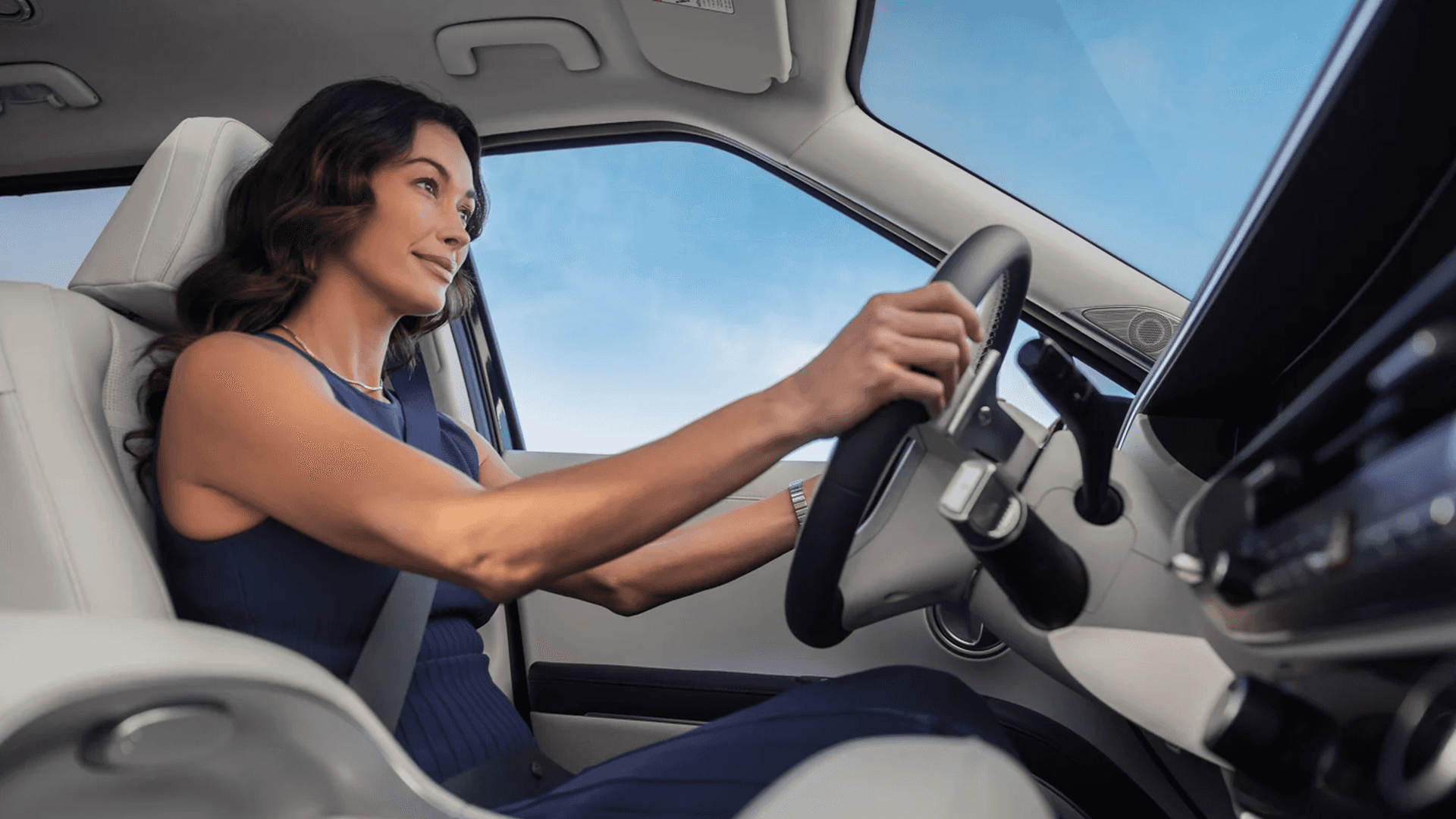Interior driver view of a woman wearing a seat belt steering a vehicle, with light colored seats, a wide windshield and blue sky, and a partial view of the dashboard and infotainment area.