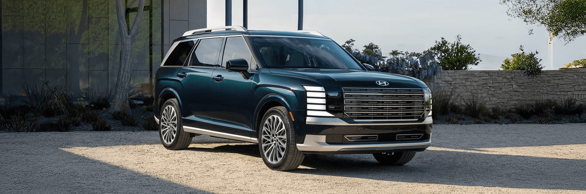 Front three quarter view of a dark green 2026 Hyundai Palisade parked on a gravel drive outside a modern home, showing the wide grille, stacked LED lights, roof rails, and multi spoke wheels.
