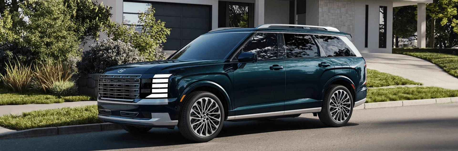 Dark green 2026 Hyundai Palisade SUV parked on a residential street in front of a modern home, shown in a front three quarter view with a wide grille, bright stacked LED lighting signature, silver lower bumper and rocker trim, large multi spoke wheels, and landscaping with trees and shrubs in the background.