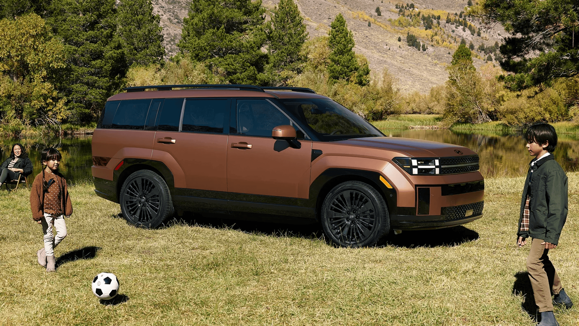 Bronze 2026 Hyundai Santa Fe parked on grass beside a lake with mountains and trees behind it, showing the boxy SUV shape, black roof rails, and dark wheels while children play nearby.