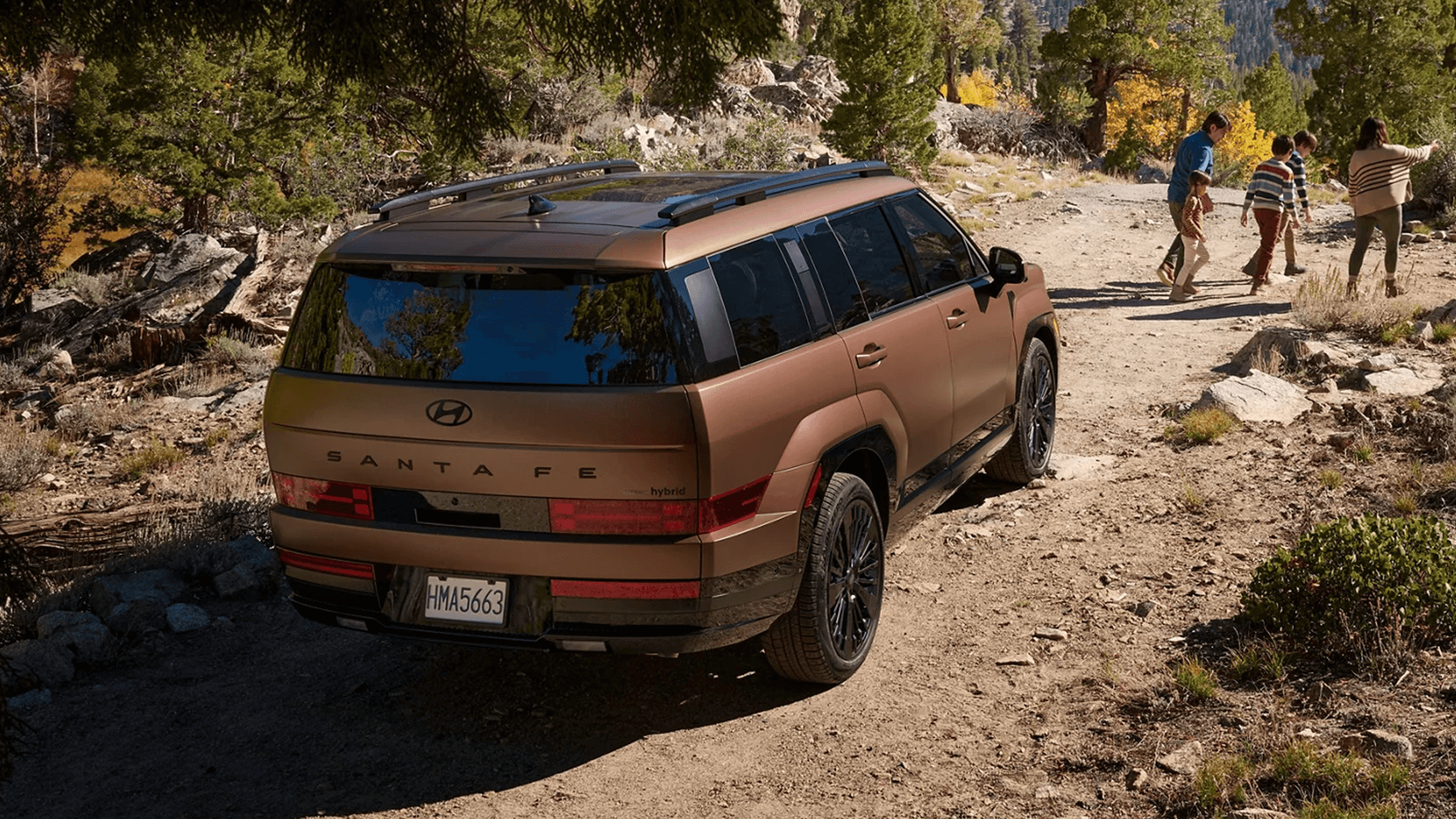 Rear three quarter view of a bronze 2026 Hyundai Santa Fe Hybrid driving on a rocky dirt road, showing the squared rear design, Santa Fe lettering, roof rails, and dark wheels as a family walks ahead.