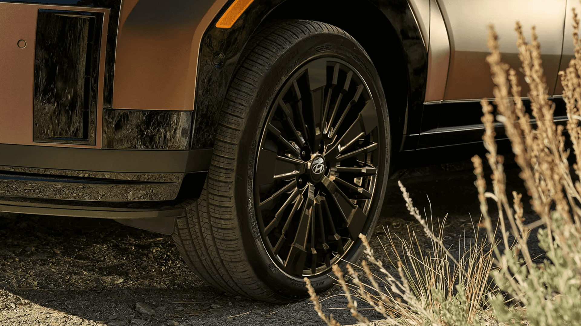 Close up of the front wheel and tire on a bronze 2026 Hyundai Santa Fe, showing a black multi spoke alloy wheel with the Hyundai center cap and the lower bumper and fender trim nearby.
