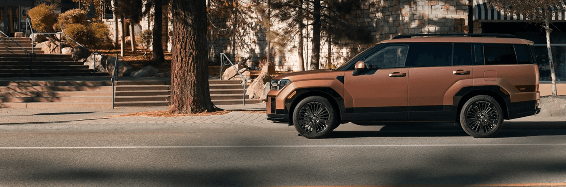 Side profile of a bronze 2026 Hyundai Santa Fe parked on a street near stone buildings and steps, showing black roof rails, dark window trim, and multi spoke wheels.