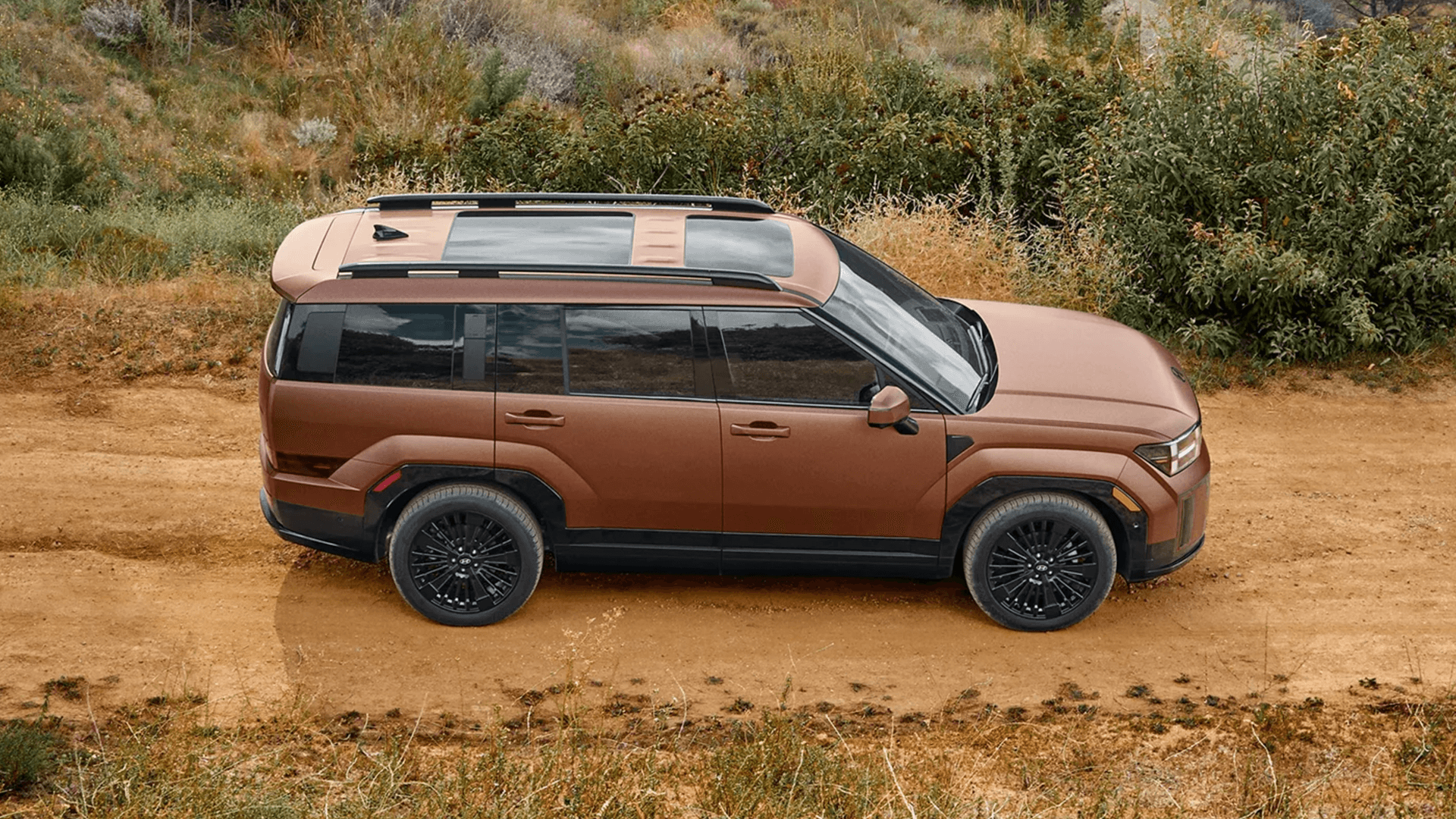 Side profile of a bronze 2026 Hyundai Santa Fe Hybrid on a dirt road, showing black wheels, squared body lines, roof rails, tinted windows, and a panoramic style roof panel, with brush and greenery behind it.