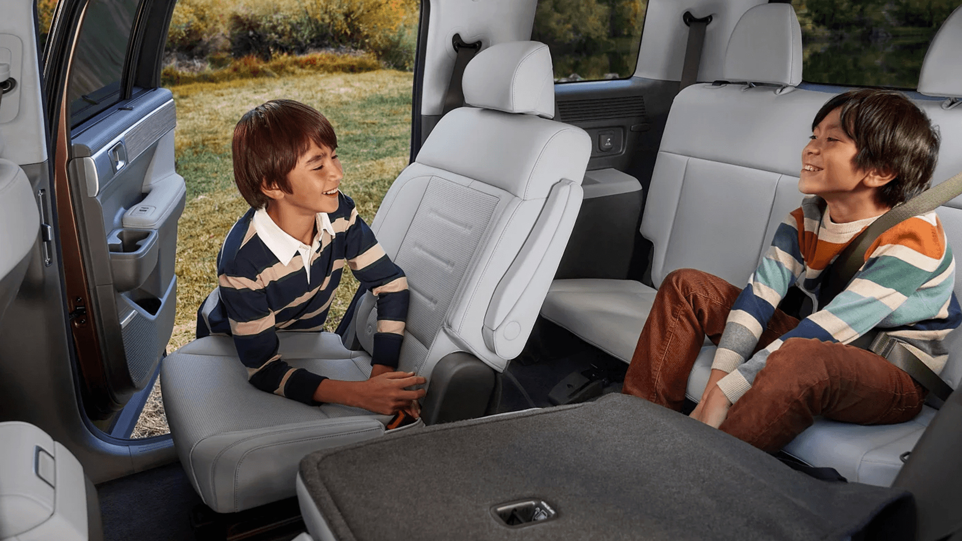 Rear cabin view of a 2026 Hyundai Santa Fe Hybrid with light gray seating, two kids smiling and talking, and a folded seatback creating a flat cargo surface in the foreground, with a side door open to a grassy outdoor setting.
