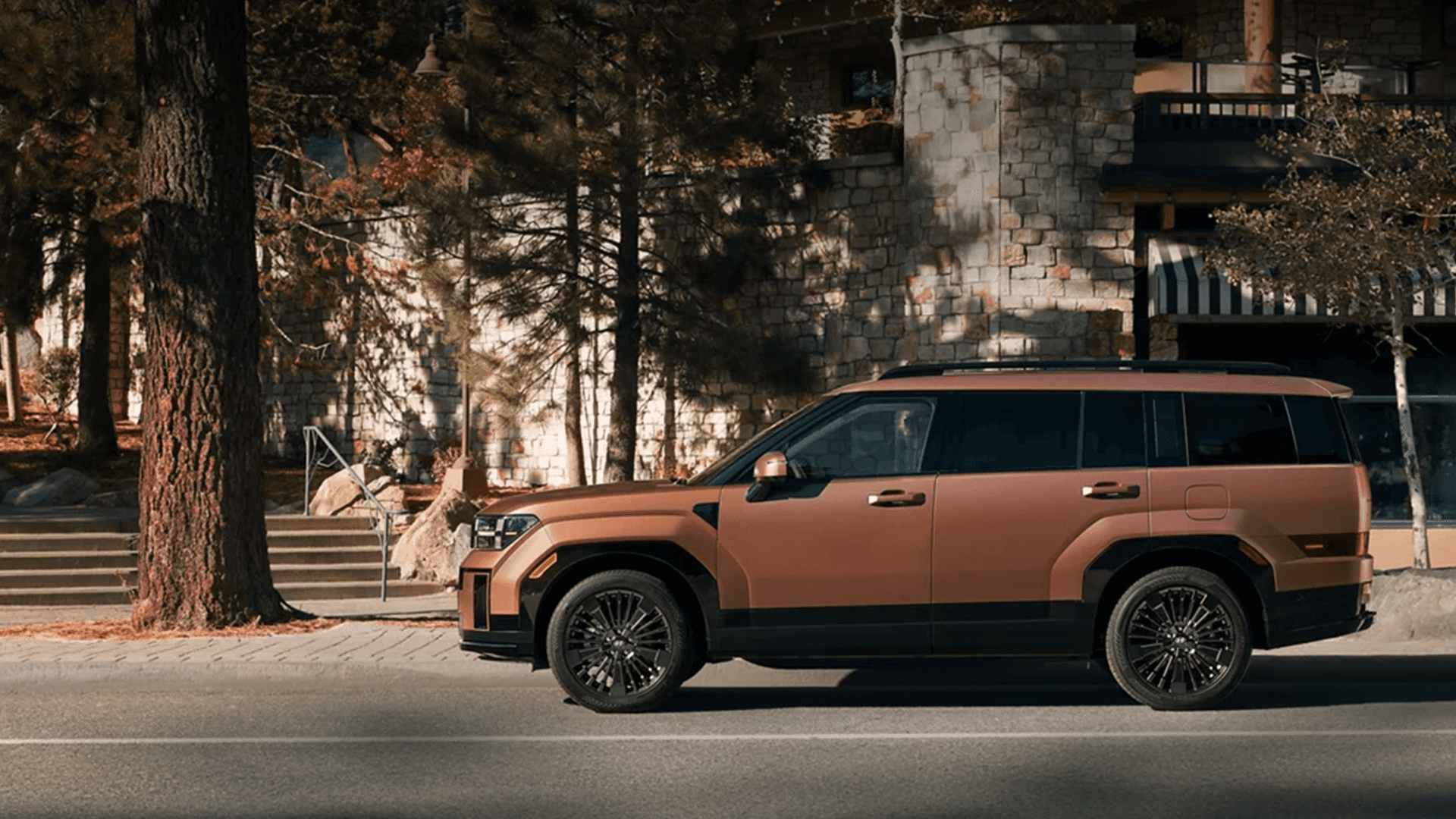 Bronze 2026 Hyundai Santa Fe Hybrid parked in profile along a street, featuring black wheels and dark window trim, with tall trees and a stone building in the background.