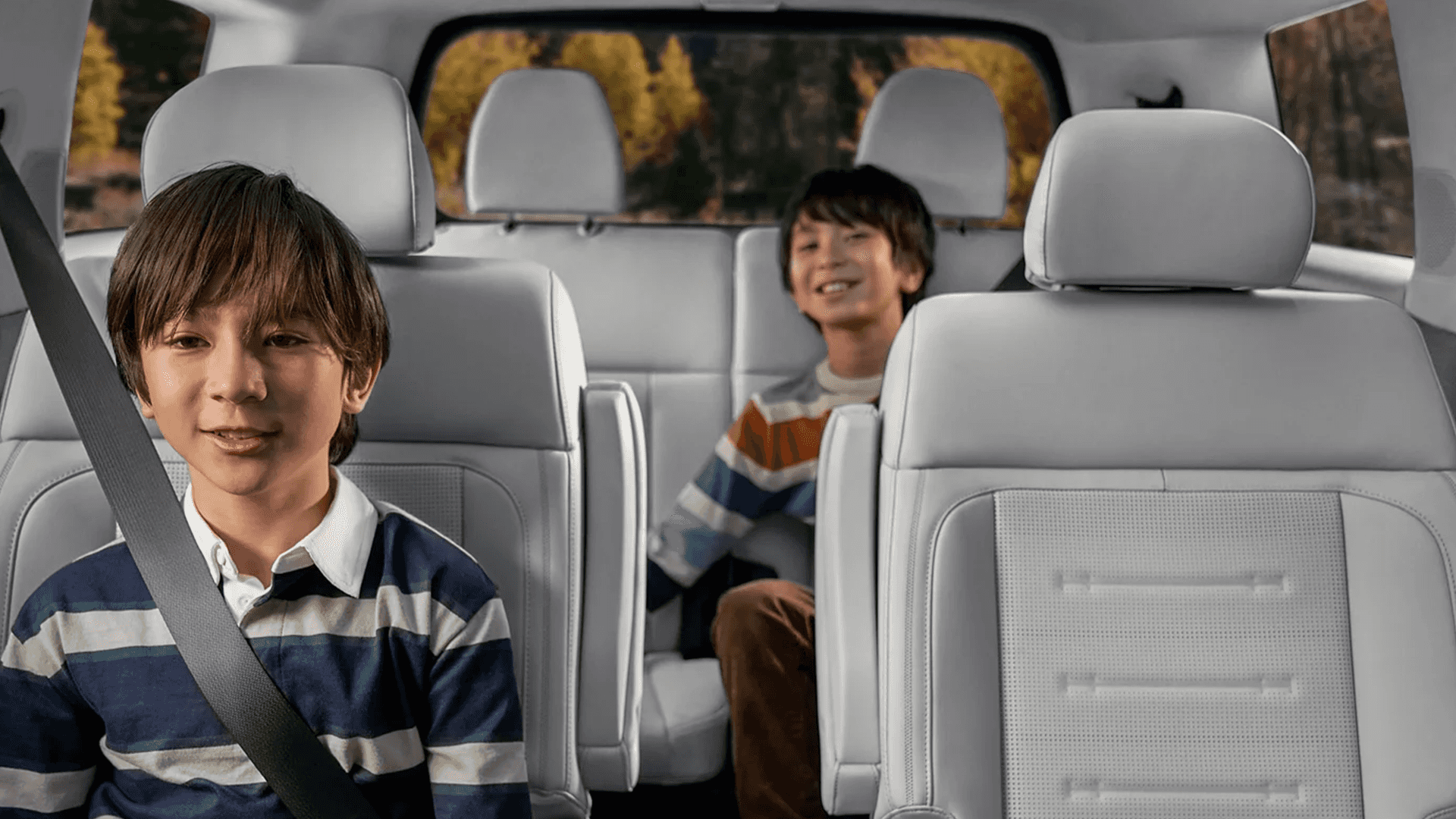 Third row and second row view inside the 2026 Hyundai Santa Fe Hybrid showing light colored seats, wide headrests, and two kids seated across the cabin with the rear window framing a wooded fall landscape.