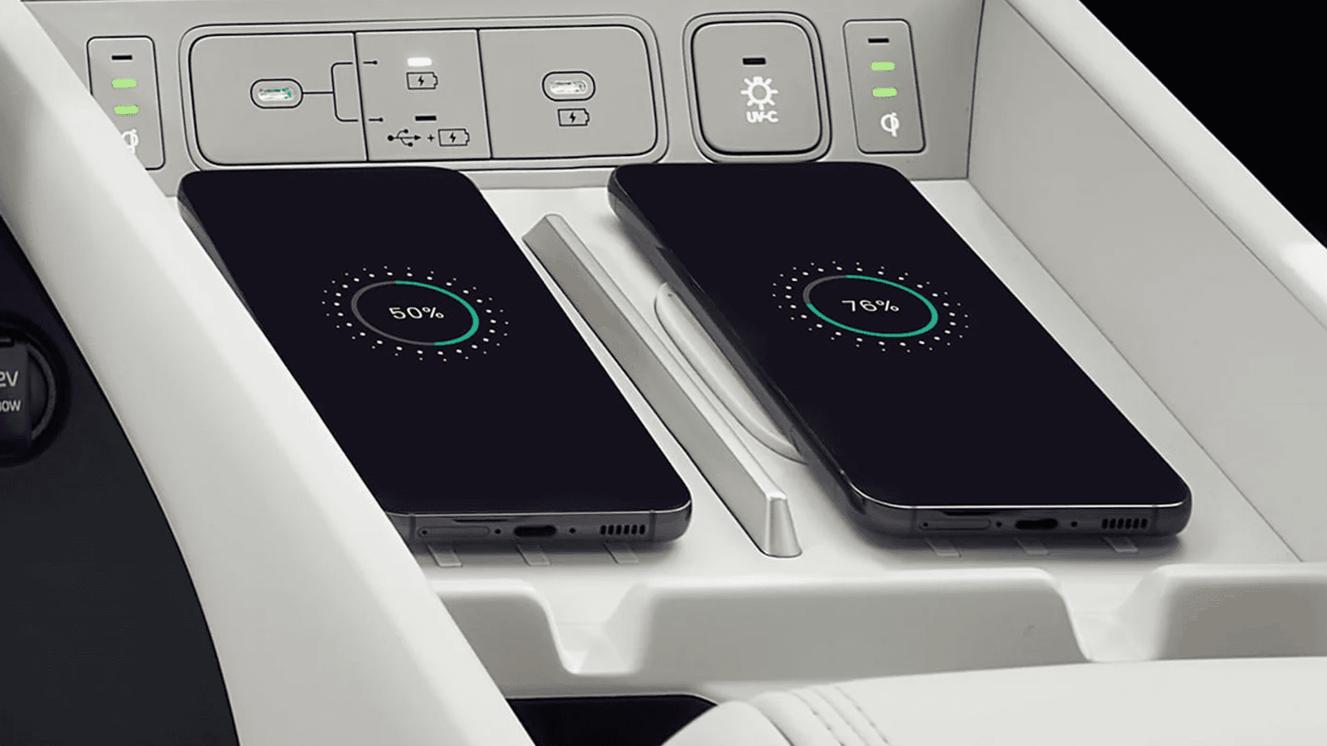 Close up of a center console wireless charging pad with two smartphones charging side by side, showing battery percentage rings, with nearby USB ports and control buttons above the tray.