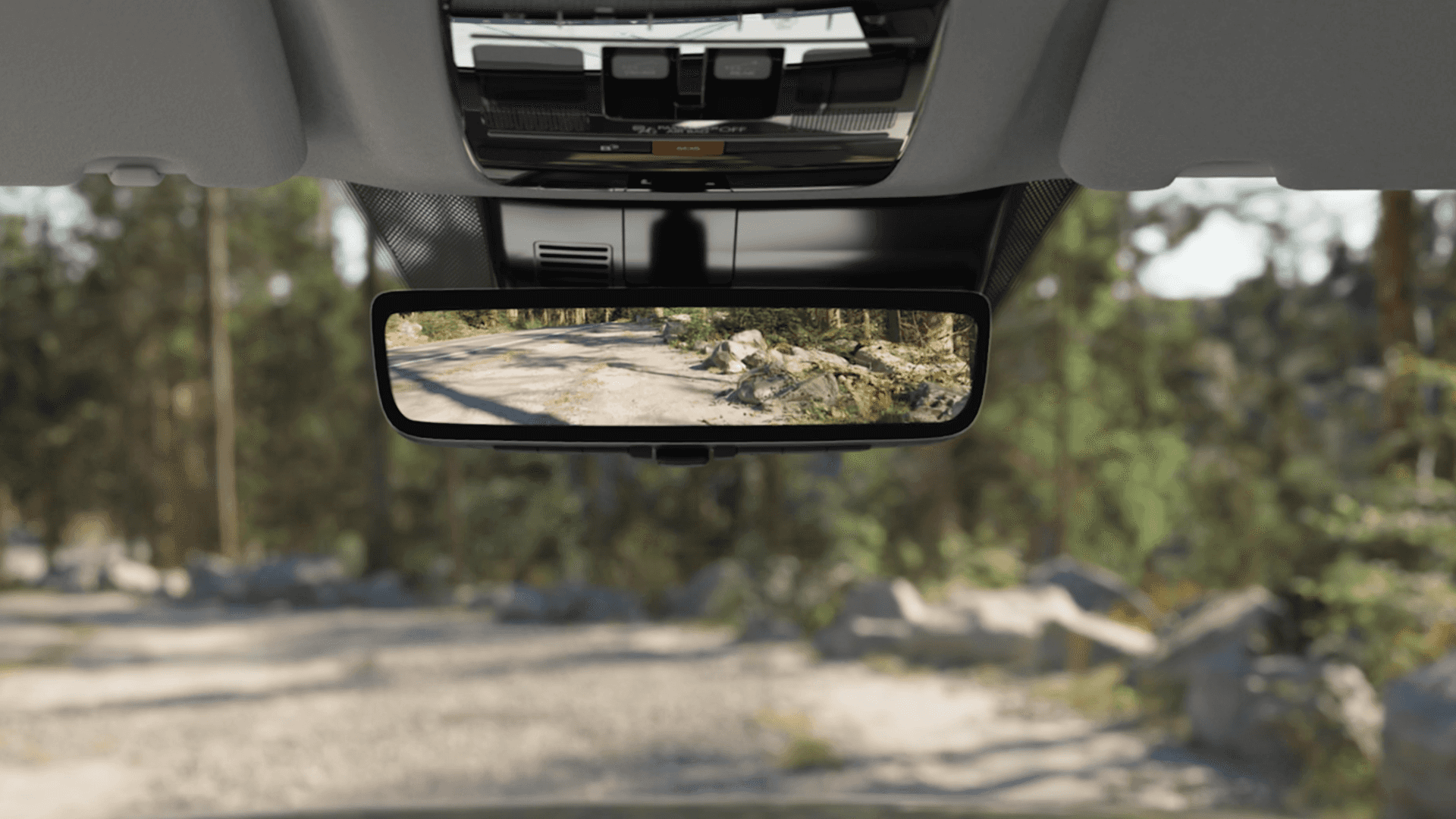 Interior close up of a digital rearview mirror displaying a live camera view of a dirt road and trees behind the vehicle, with the mirror mounted below the overhead console.