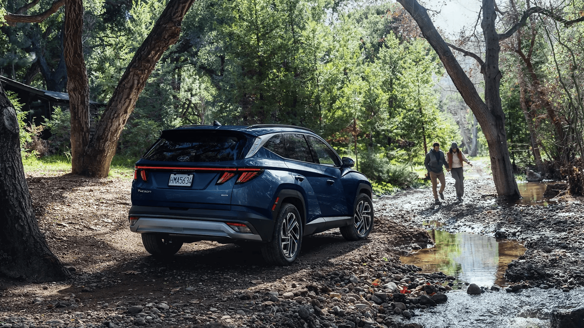 Rear three-quarter view of a blue 2026 Hyundai Tucson on a wooded trail near a creek, showing the rear design, taillights, roof rails, and the SUV positioned on rocky ground.
