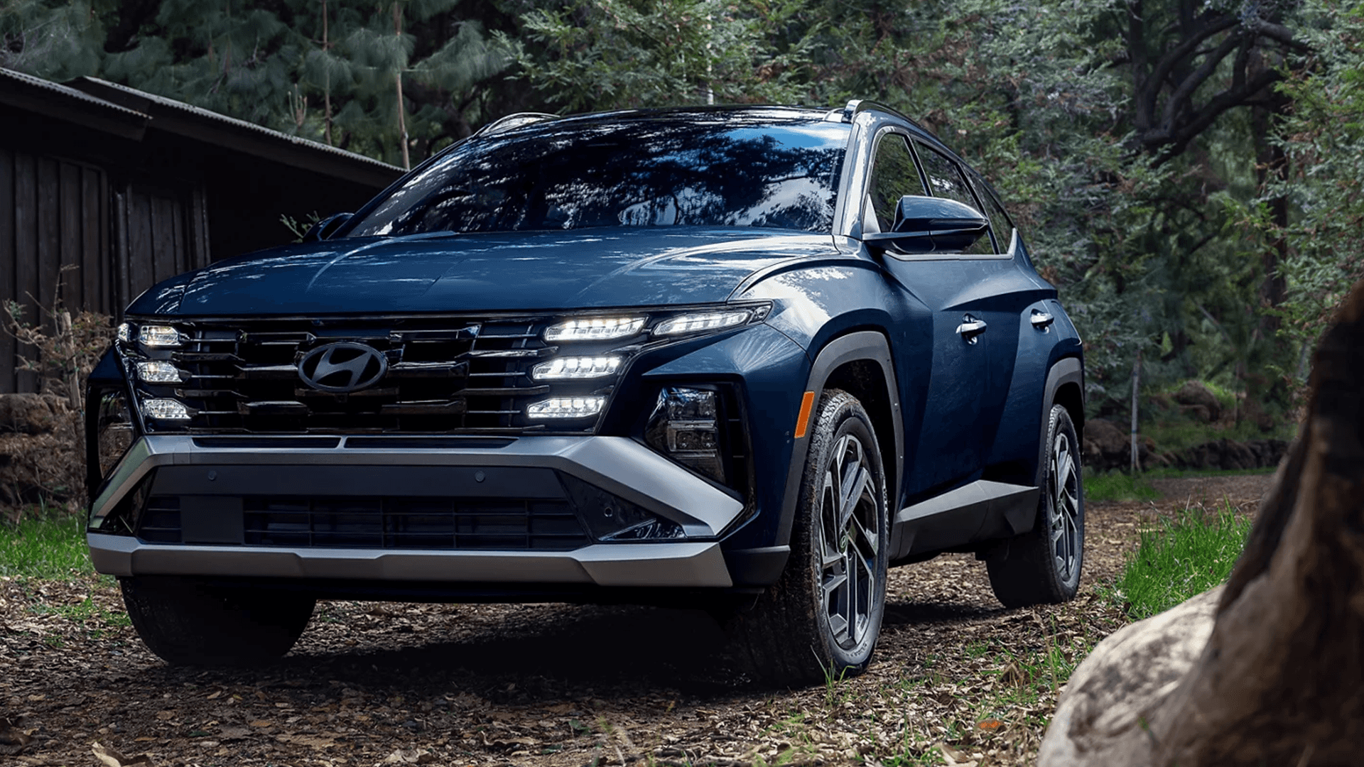 Front three-quarter view of a blue 2026 Hyundai Tucson on a forest road, with the LED lighting signature illuminated across the grille and front fascia.
