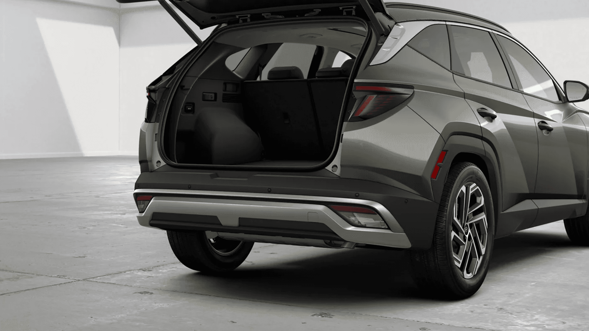 Rear three-quarter studio view of an olive green 2026 Hyundai Tucson with the liftgate open, showing the wide cargo opening, rear bumper trim, taillights, and a multi-spoke alloy wheel.