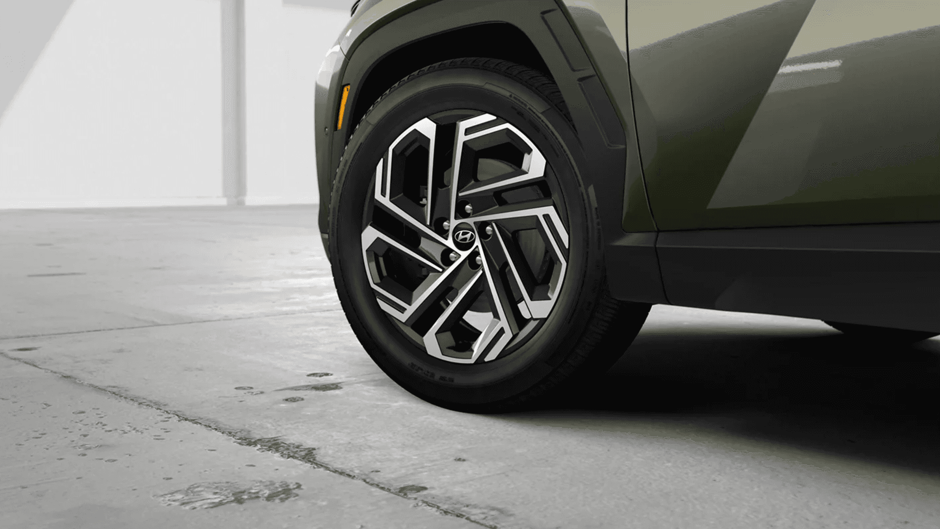 Close-up studio shot of the front wheel on an olive green 2026 Hyundai Tucson, highlighting the two-tone multi-spoke alloy wheel design, Hyundai center cap, tire sidewall, and black fender cladding.