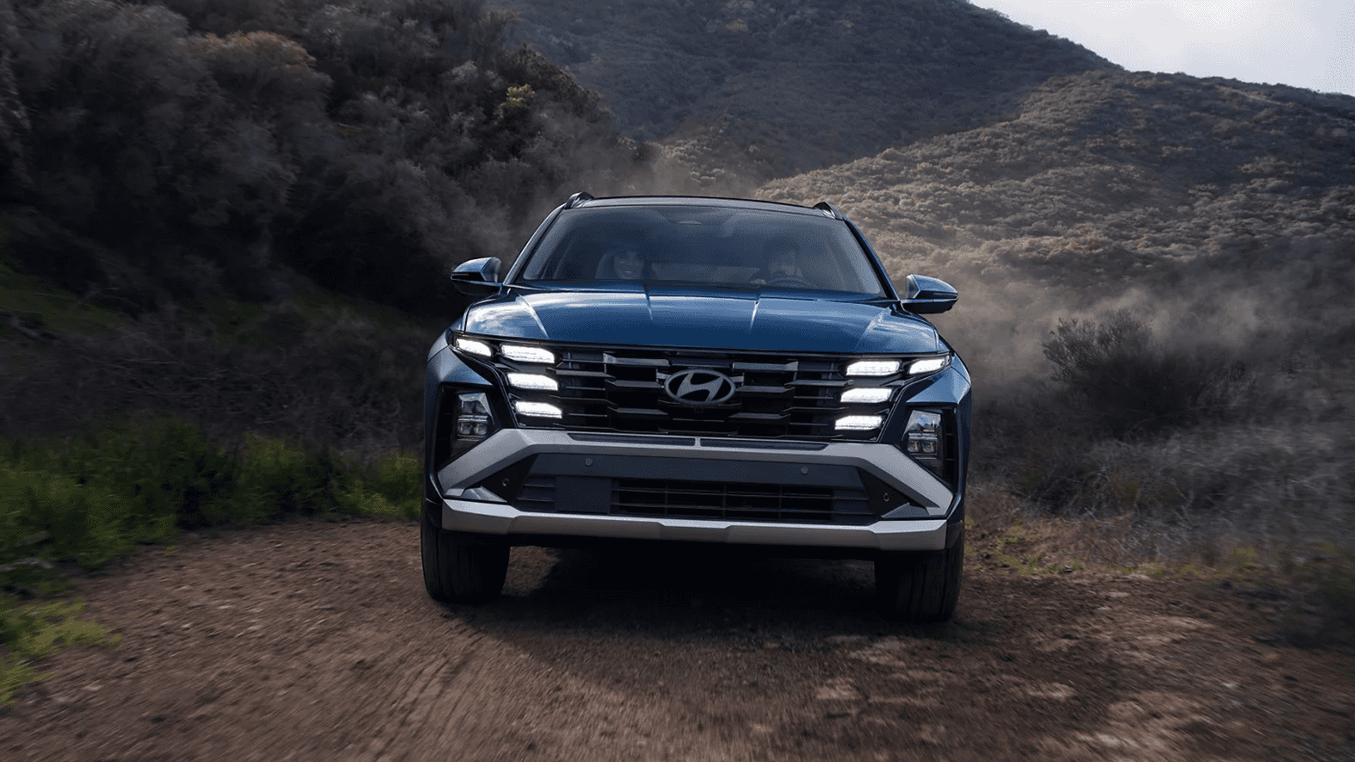 Head on view of a dark blue Hyundai Tucson driving on a dirt trail between hills, with dust in the air and the LED running lights and large grille visible from the front.