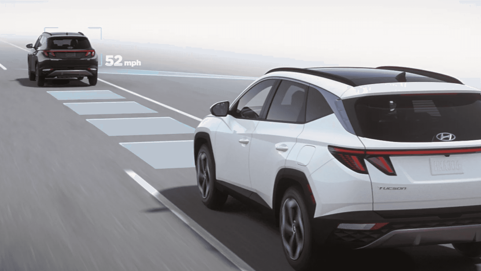Rear three quarter view of a white Hyundai Tucson on a highway with a driver assistance graphic overlay, showing blue lane and distance markers and a leading vehicle labeled 52 mph.