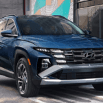 Front three quarter view of a dark blue Hyundai Tucson driving on a city street, showing the wide grille, stacked LED lighting, black cladding, and multi spoke wheels beside modern buildings.