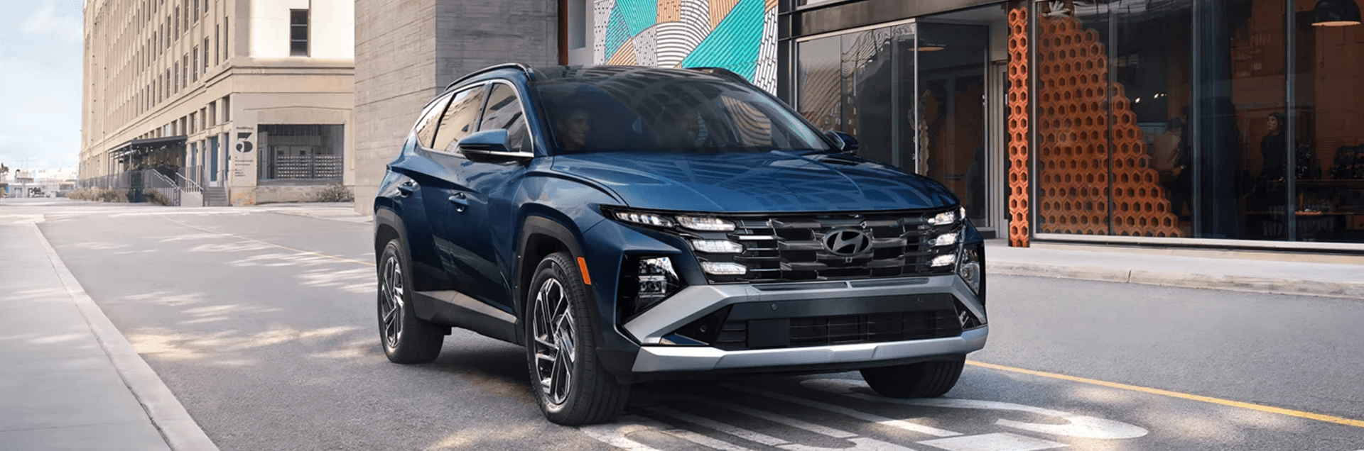 Front three quarter view of a dark blue Hyundai Tucson driving on a city street, showing the wide grille, stacked LED lighting, black cladding, and multi spoke wheels beside modern buildings.
