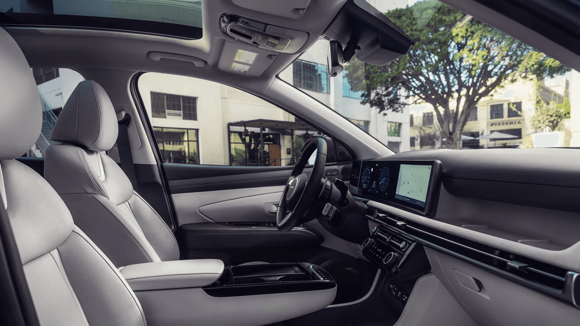 Wide interior overview from the front passenger side showing the 2026 Hyundai Tucson cabin with light gray seats, a panoramic sunroof, a wide dashboard display, center climate controls, and a glossy black center console.