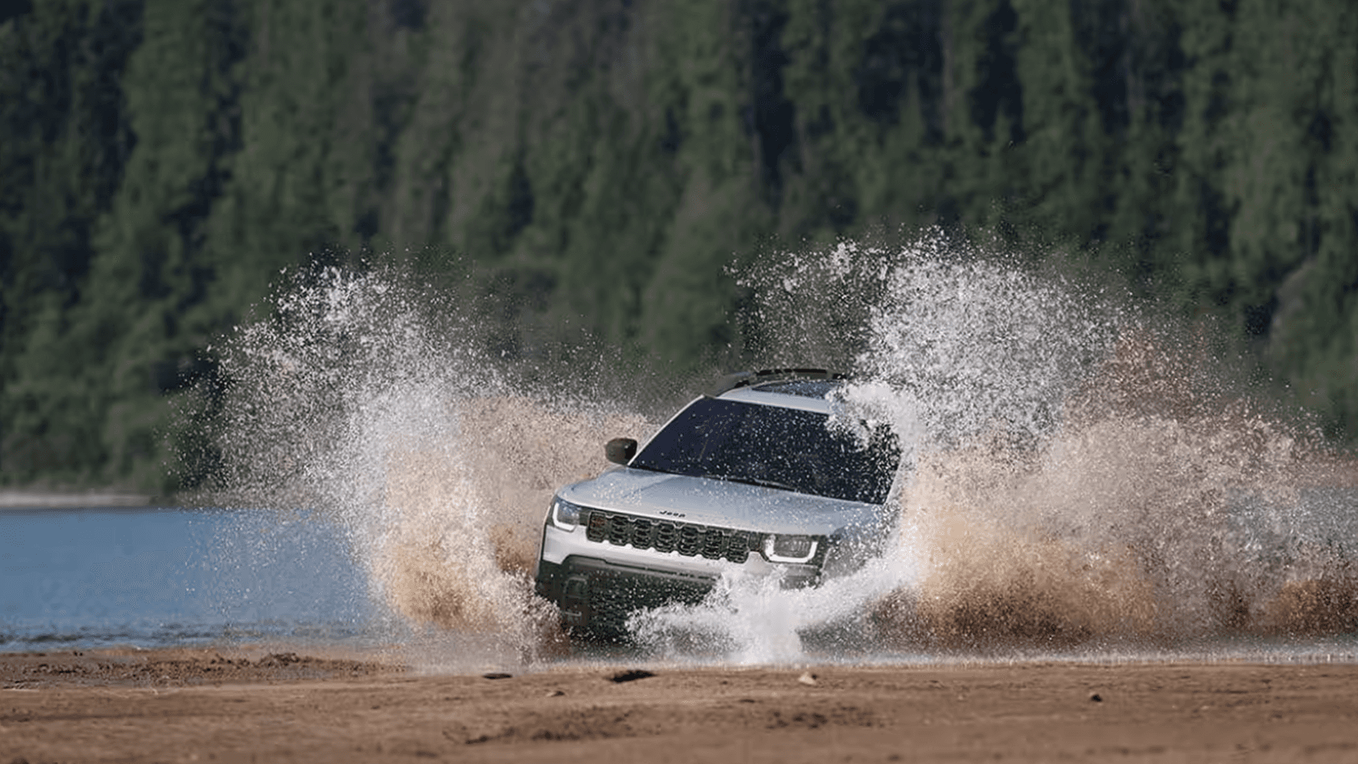 White Jeep Cherokee charging through shallow water near a forested shoreline, with large splashes around the front end and the seven-slot grille visible.