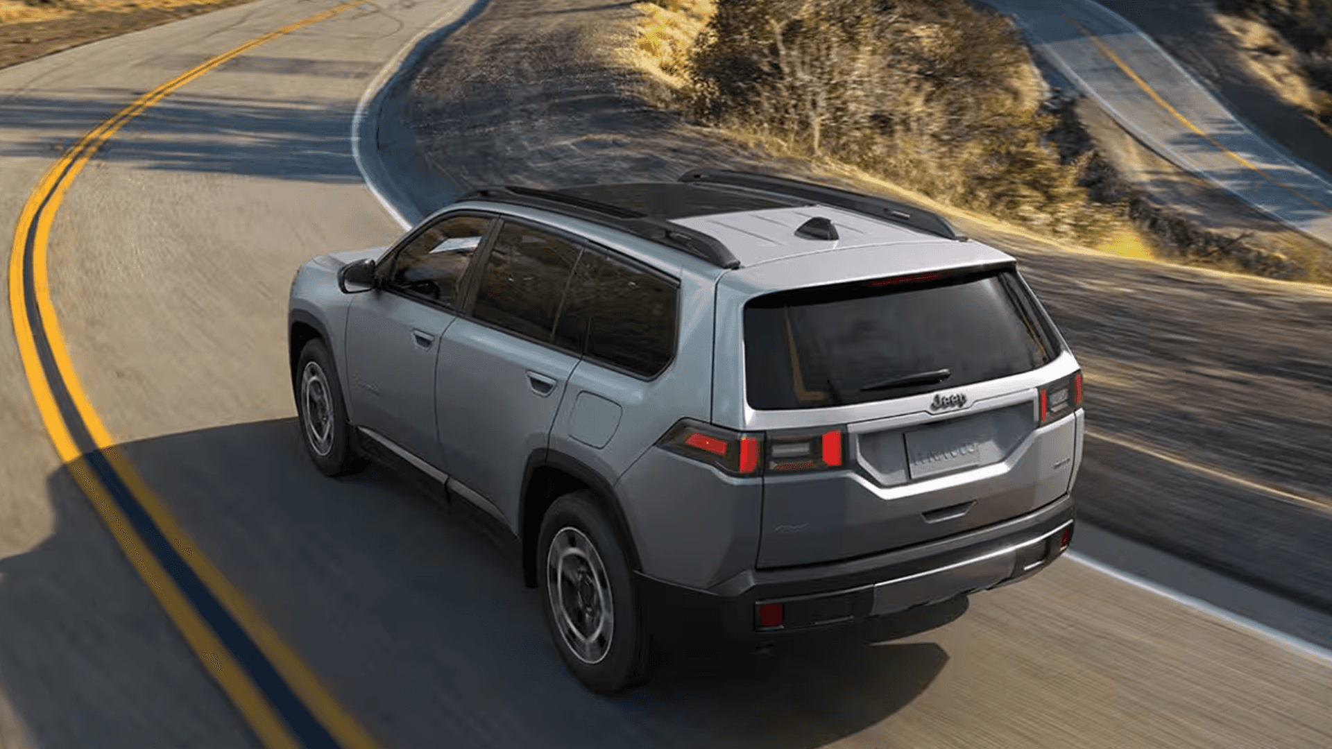 Rear three-quarter overhead view of a gray Jeep Cherokee driving on a winding two-lane road, showing roof rails, rear spoiler, taillights, and tinted rear glass.