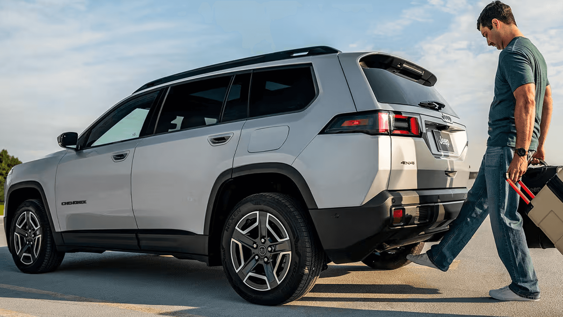 Side view of a white Jeep Cherokee parked with the rear liftgate open as a man steps near the bumper holding luggage, highlighting hands-free cargo access and 4x4 badging.