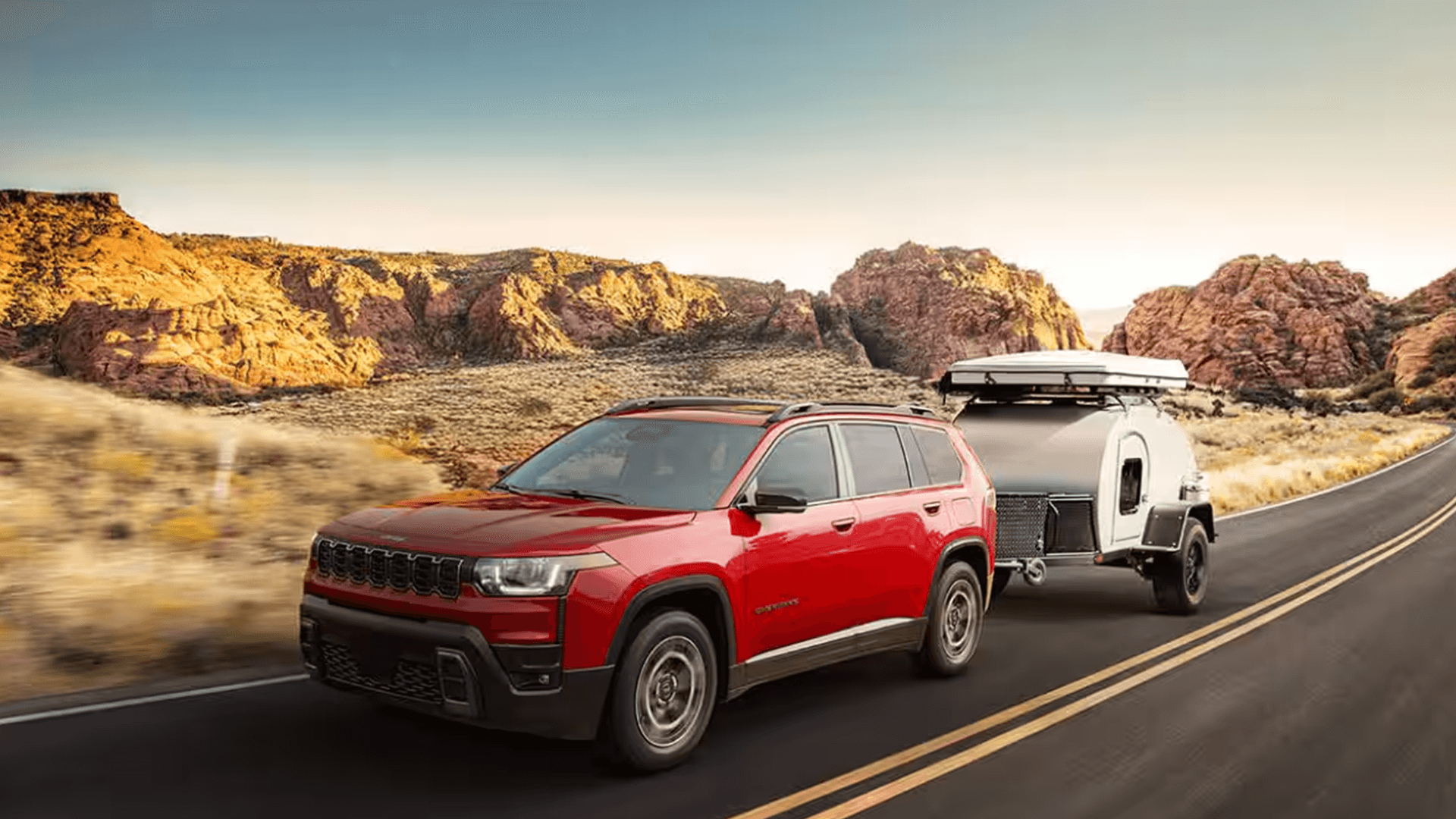 Red 2026 Jeep Cherokee towing a small camper trailer on a desert highway with rocky hills in the distance.