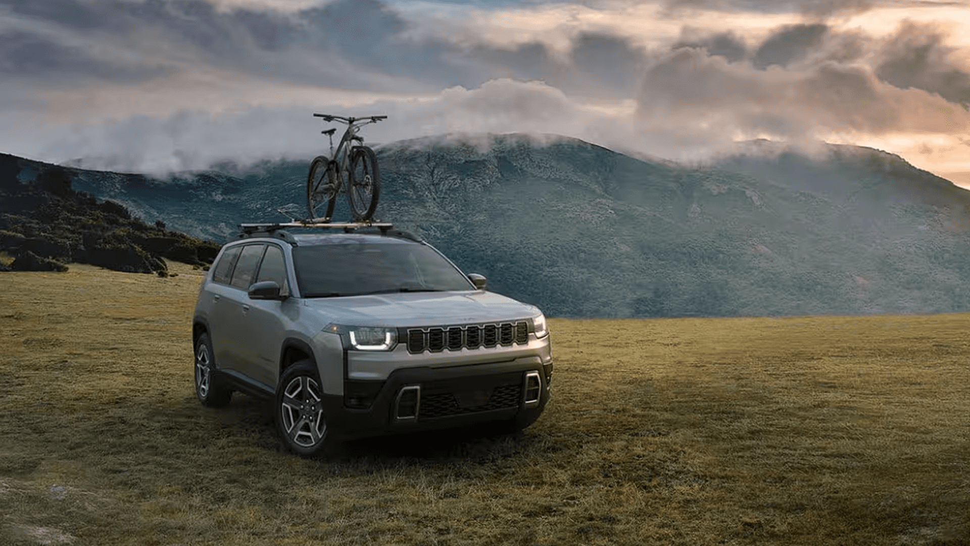 Silver 2026 Jeep Cherokee parked in a grassy field with a bicycle mounted on the roof and mountains under cloudy skies.