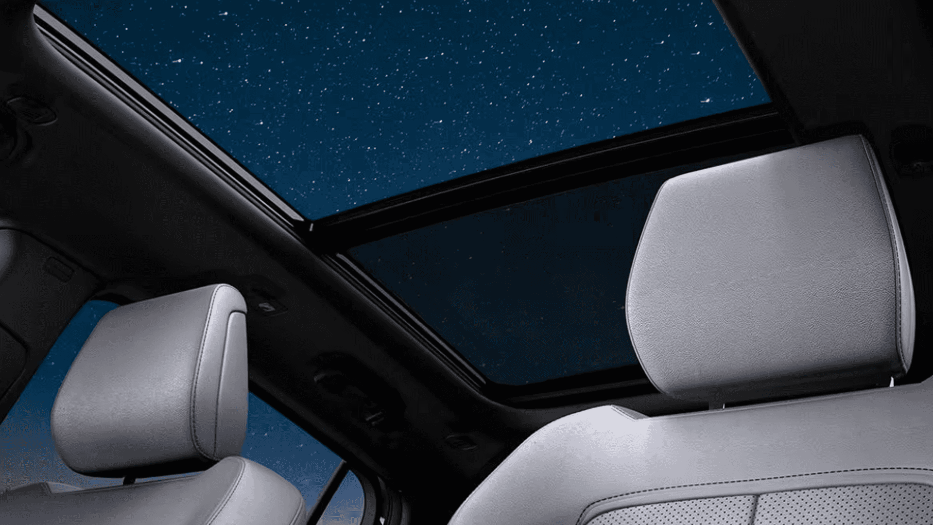 Upward interior view of the 2026 Jeep Cherokee showing light gray front seat headrests beneath a large panoramic glass roof with a starry night sky visible overhead.
