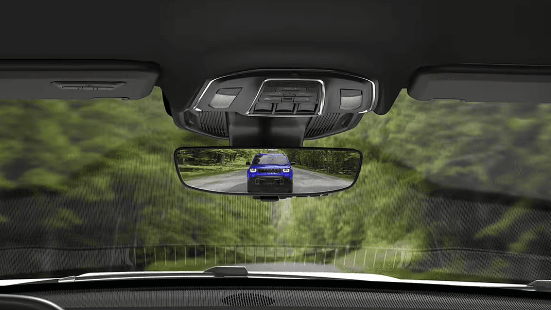 Interior view toward the windshield showing the overhead console and rearview mirror, with the mirror reflecting a blue SUV following on a two lane road.
