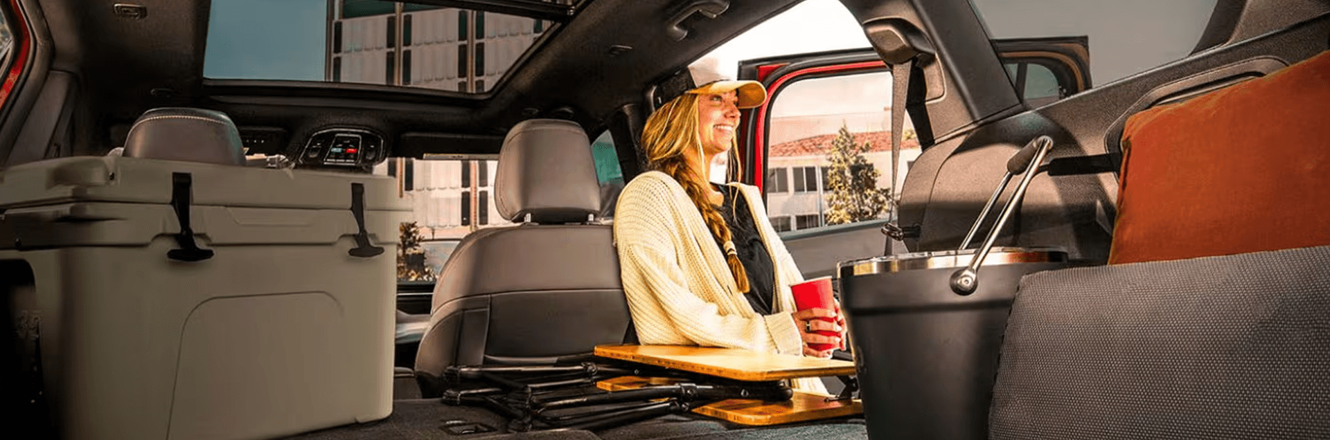 Rear cabin view of the 2026 Jeep Cherokee with the second row folded flat, showing a cooler, folded camp chairs, and gear on the cargo floor under a panoramic glass roof.