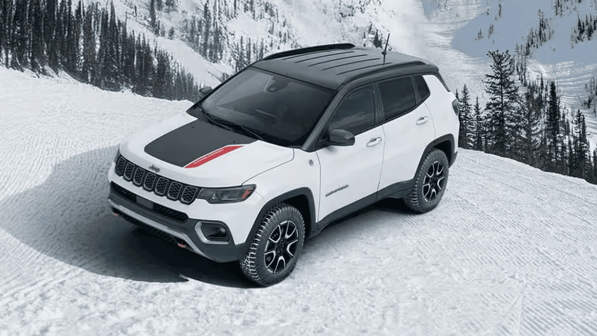 White 2026 Jeep Compass Trailhawk on a snowy mountain road, viewed from above, showing black roof, black hood decal with red stripe, and all-terrain tires.