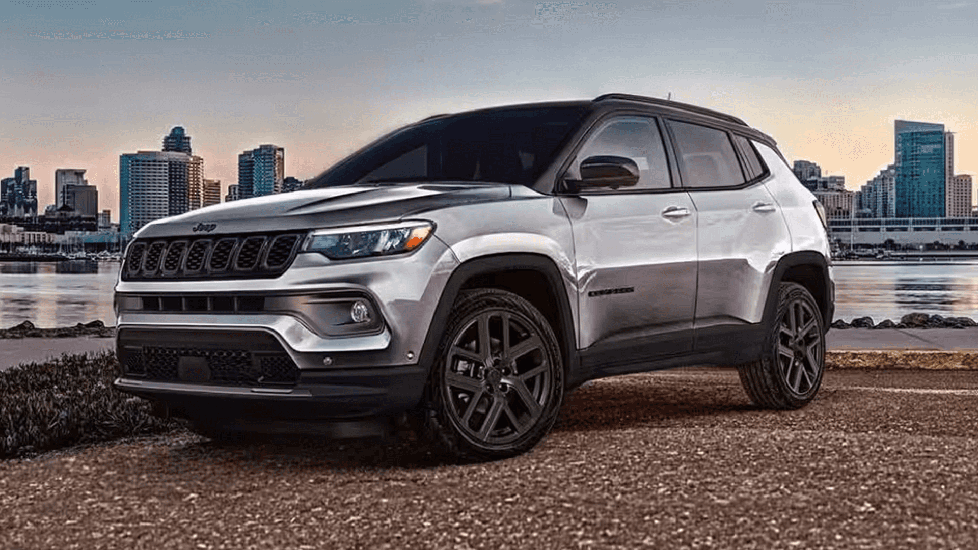 Silver 2026 Jeep Compass parked near a waterfront skyline at sunset, showing the front three-quarter view, black roof, black hood decal, and dark multi-spoke wheels.