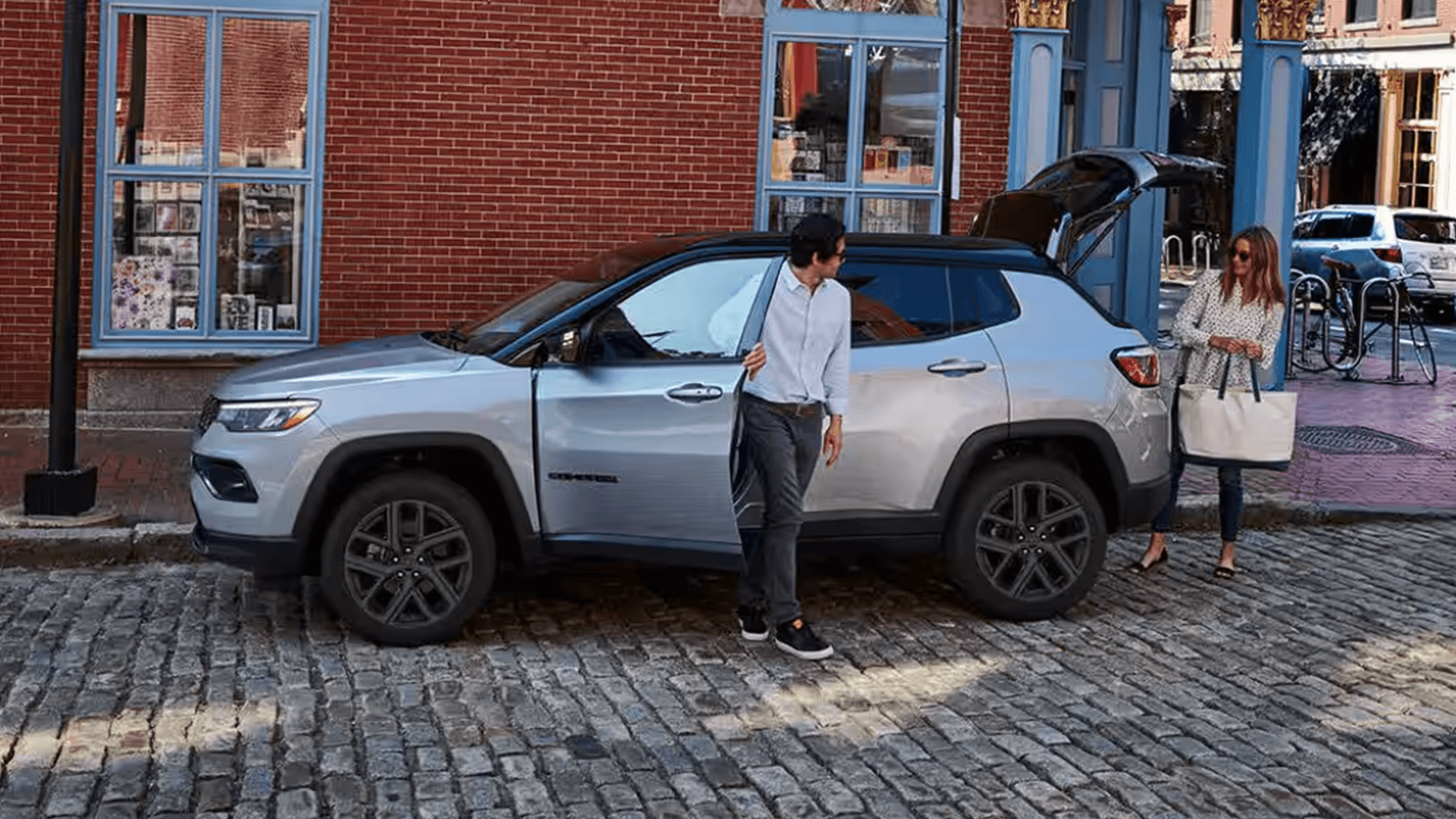 Silver 2026 Jeep Compass parked on a cobblestone street with the rear liftgate open, showing cargo access with a man by the driver door and a woman holding a tote bag nearby.